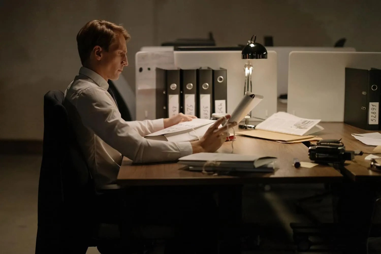 A compliance officer reads a bound filing at a desk under a lamp at night, numbered ring binders lined up behind him.