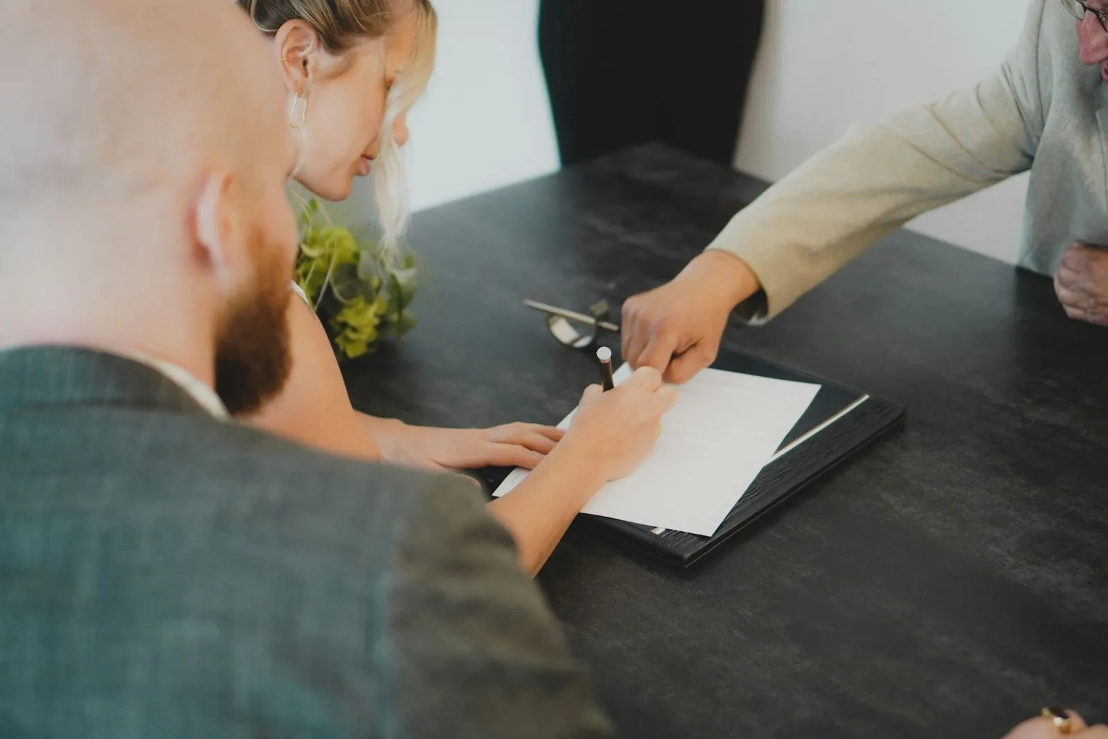 Three colleagues at a dark meeting table reviewing a document in a folder; one holds a pen over the page while another points to a line on it.